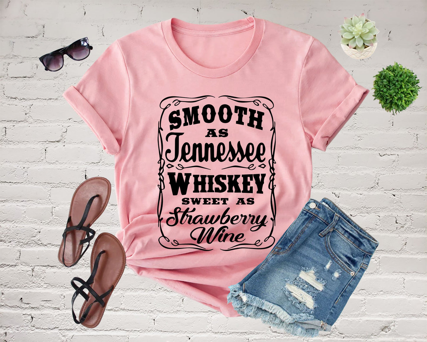 Pink t-shirt with text, sunglasses, denim shorts, sandals, and plants.