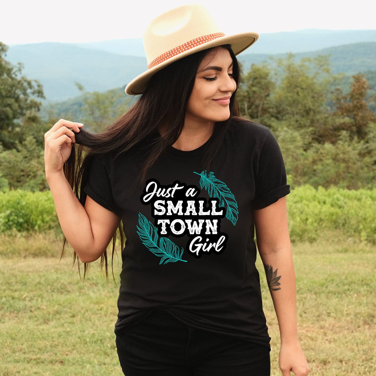 Woman in Small Town Girl tee, hat, outdoors.