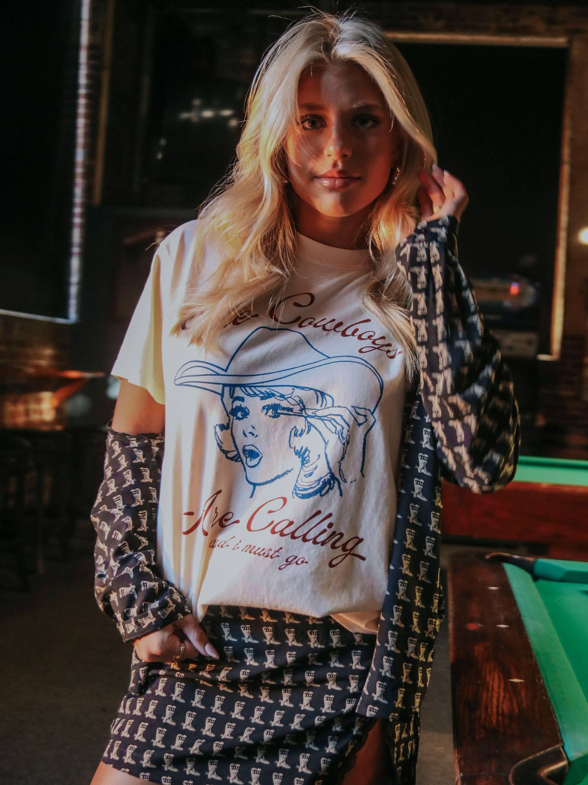 Blonde woman in t-shirt, skirt, and jacket, near a pool table.