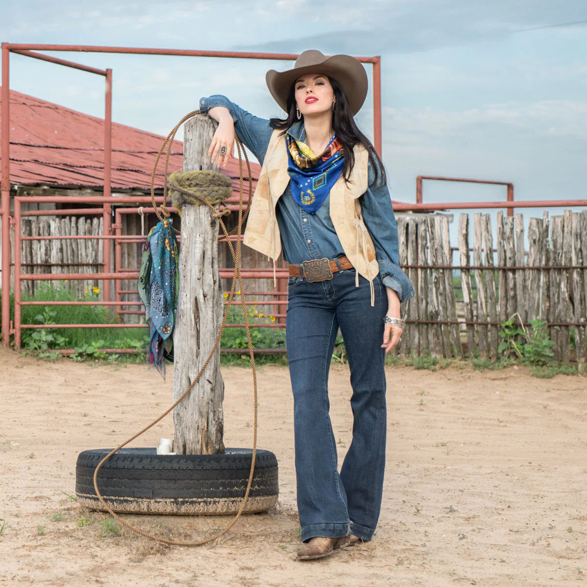 Cowgirl in royal blue jeans and vintage horse scarf, leaning on post.