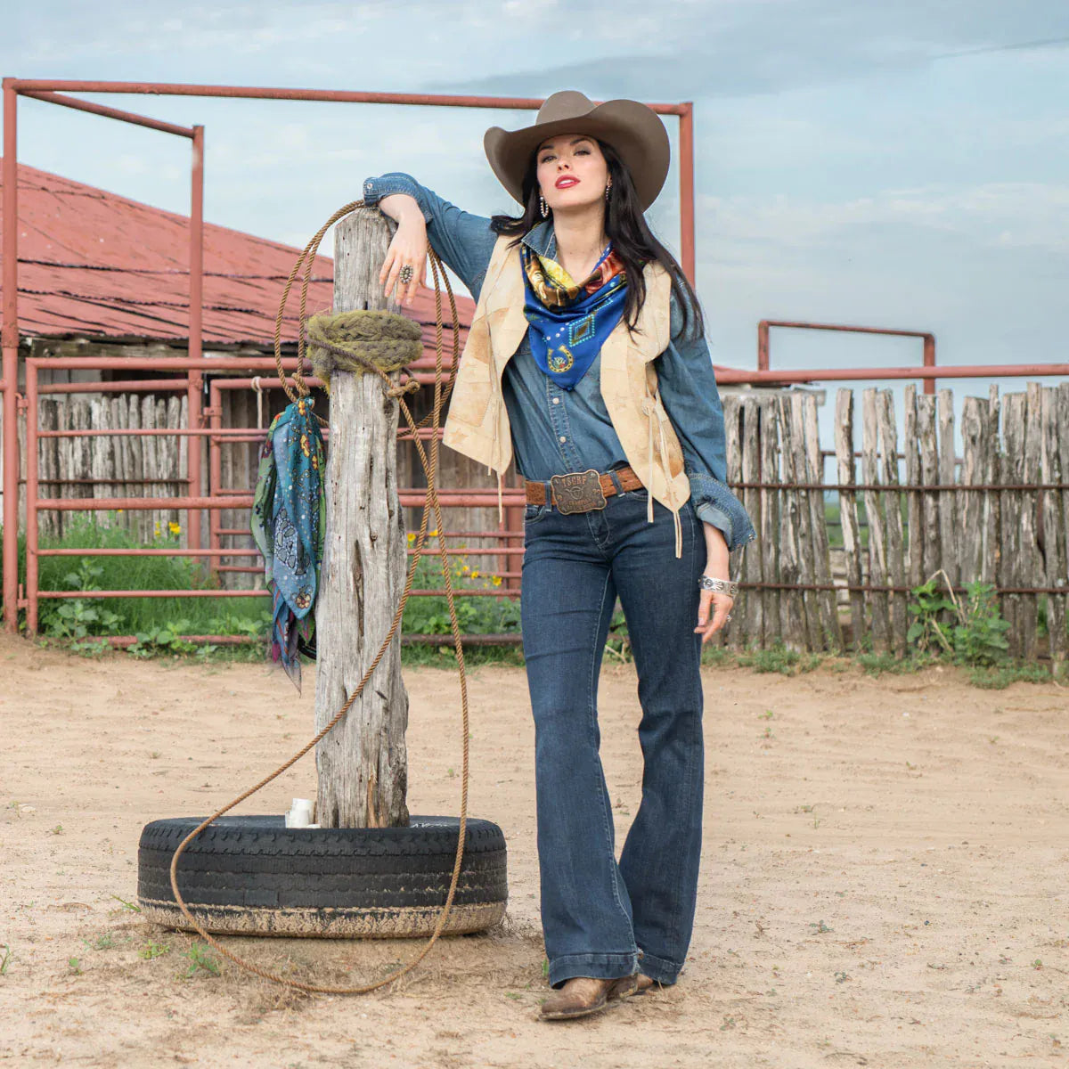 Cowgirl in royal blue jeans and vintage horse scarf, leaning on post.